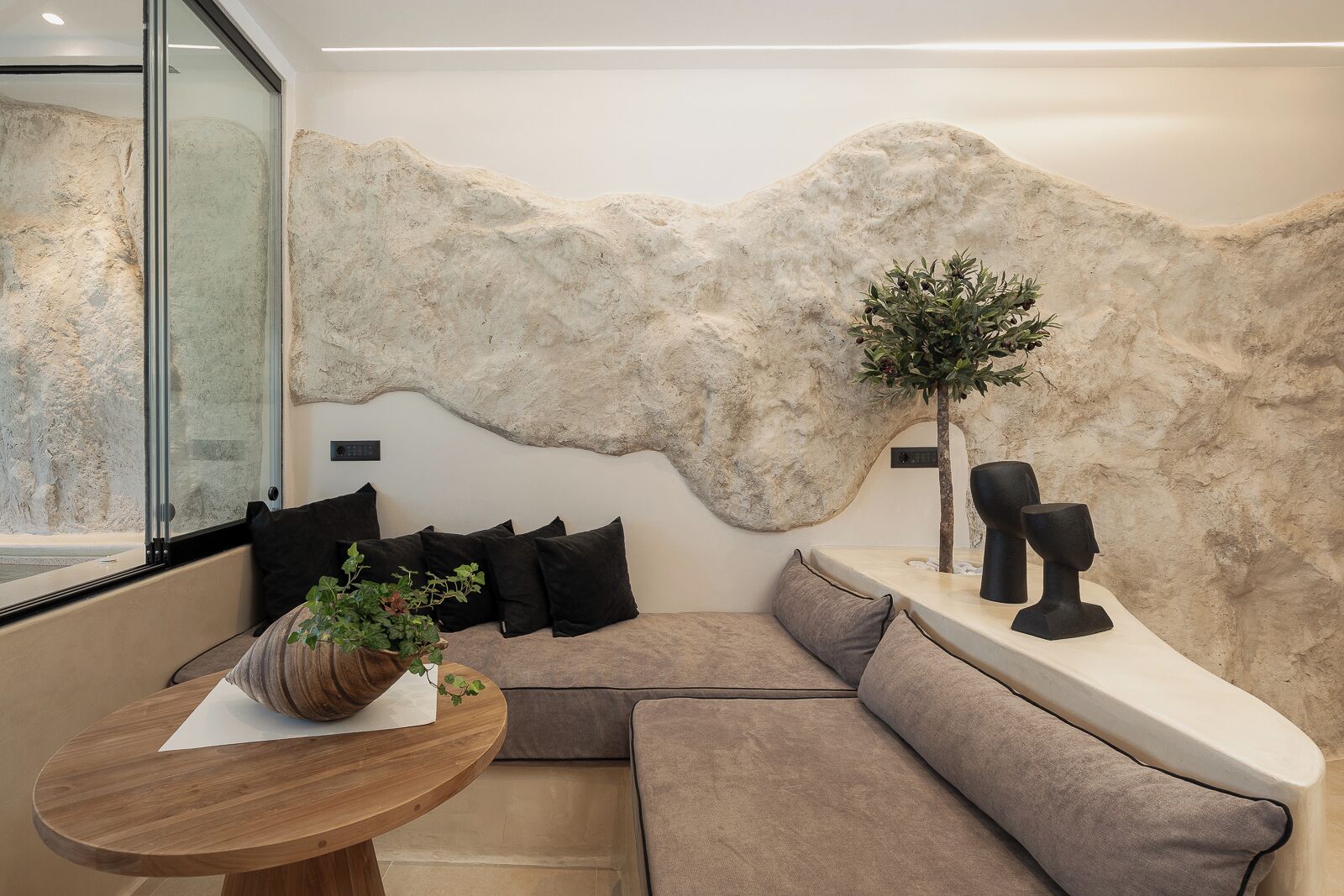 Ein modernes Interieur mit einer Steinsäule, einer Sitzecke mit Kissen, einem Holztisch mit Pflanze und einem kleinen Baum.