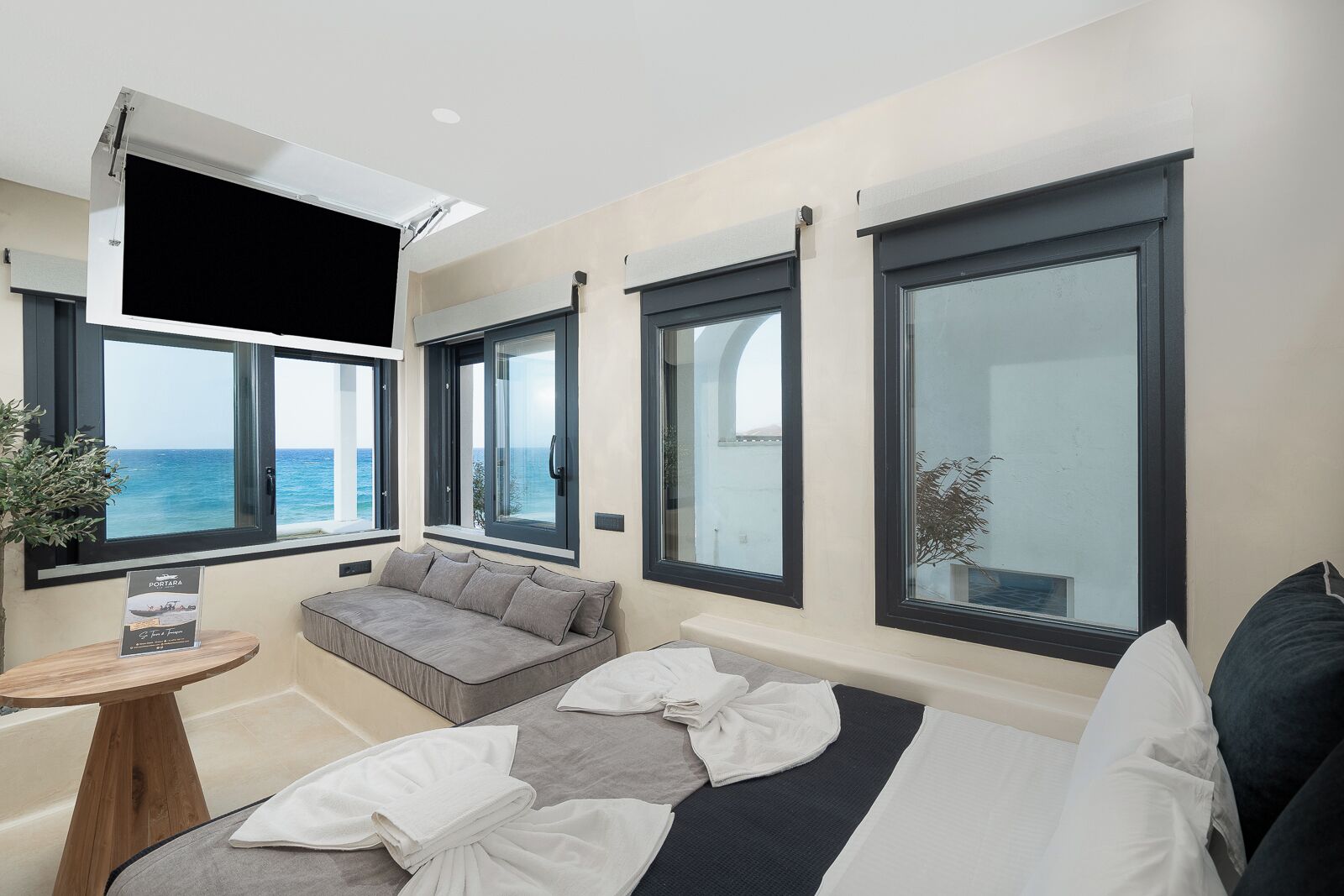 Habitación de hotel moderna con una cama grande, un sofá y una mesita. Varias ventanas ofrecen vistas al mar.