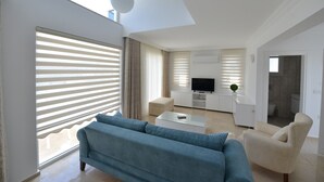 A modern living room with a sofa, coffee table, and TV, large windows with blinds, and a view into the kitchen.