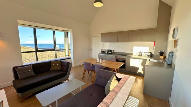 Un intérieur moderne comprenant un canapé, une table à manger, des chaises et un coin cuisine avec vue sur la plage.