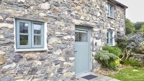 A stone building with a grey door and windows, surrounded by a garden with shrubs and a stone wall.