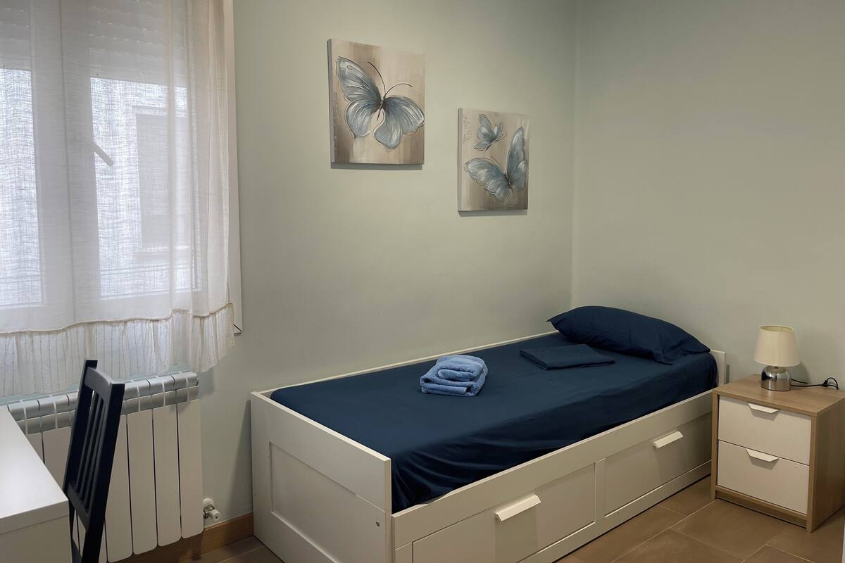 A bedroom with a bed, a nightstand, a chair, and two butterfly paintings on the wall.