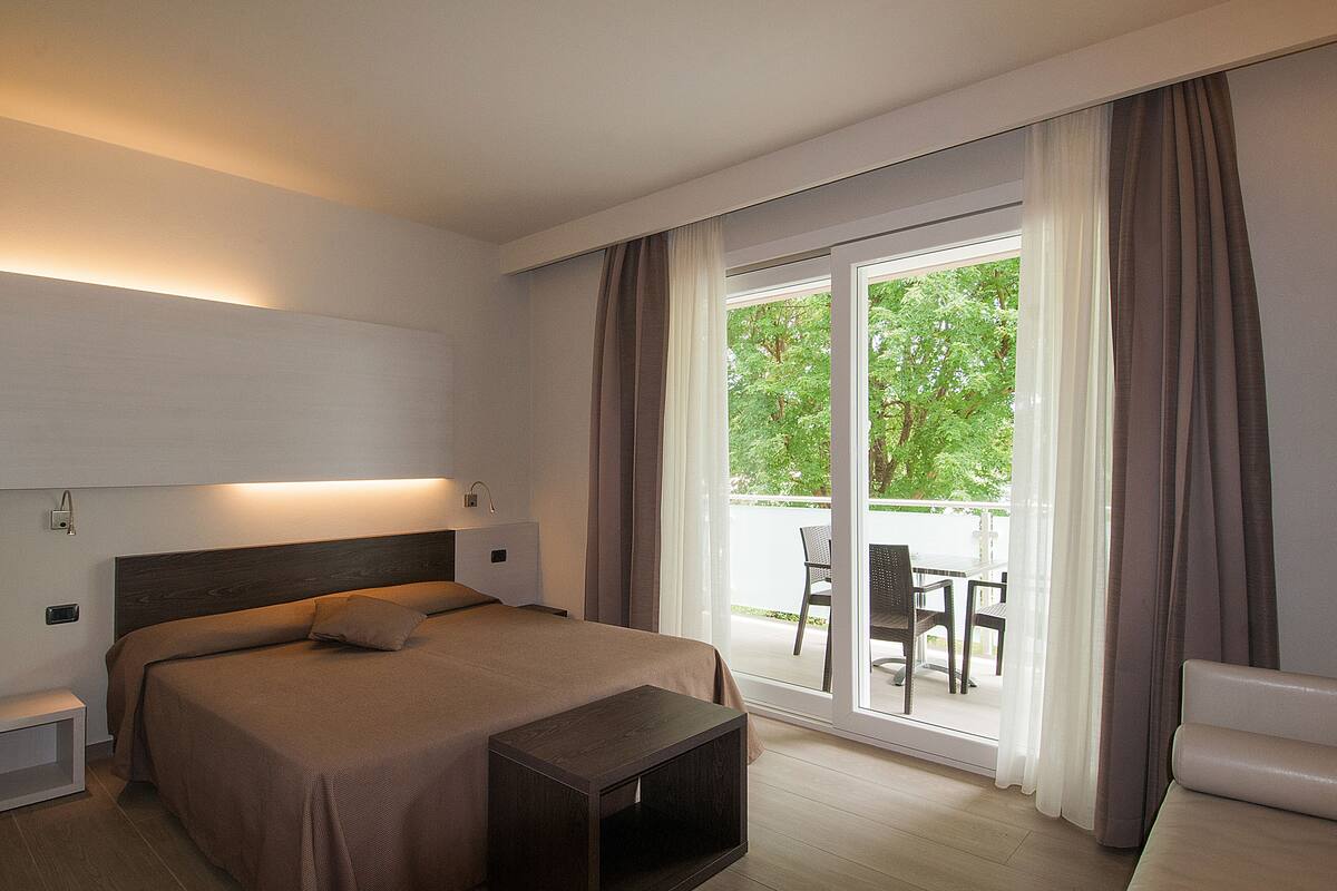 A modern hotel room with a large bed, a bedside table, a small wooden nightstand, and a view of the outdoors through a sliding glass door.