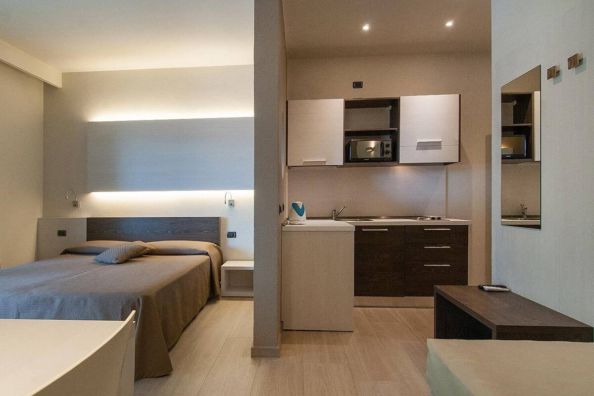 A compact hotel room with a bed, bedside table, and a kitchenette area including a microwave and sink.
