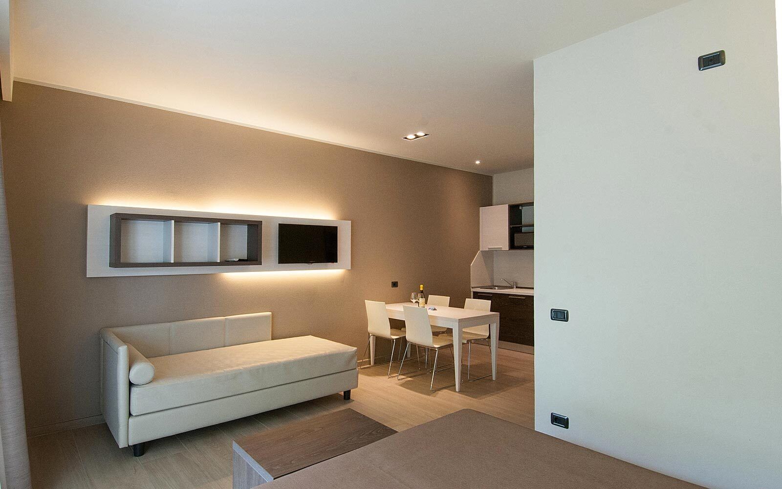 A modern living room with a sofa, a dining table with chairs, a TV mounted on the wall, and a kitchen area visible through an open door.