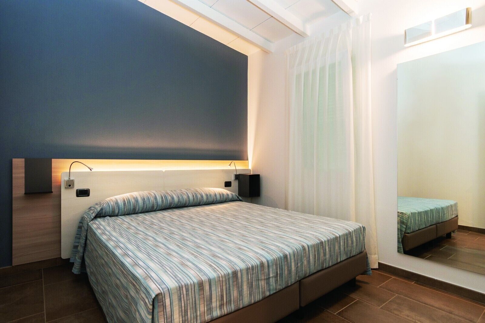 Un dormitorio moderno con cama, lámparas de noche, un espejo grande y una ventana con cortinas.