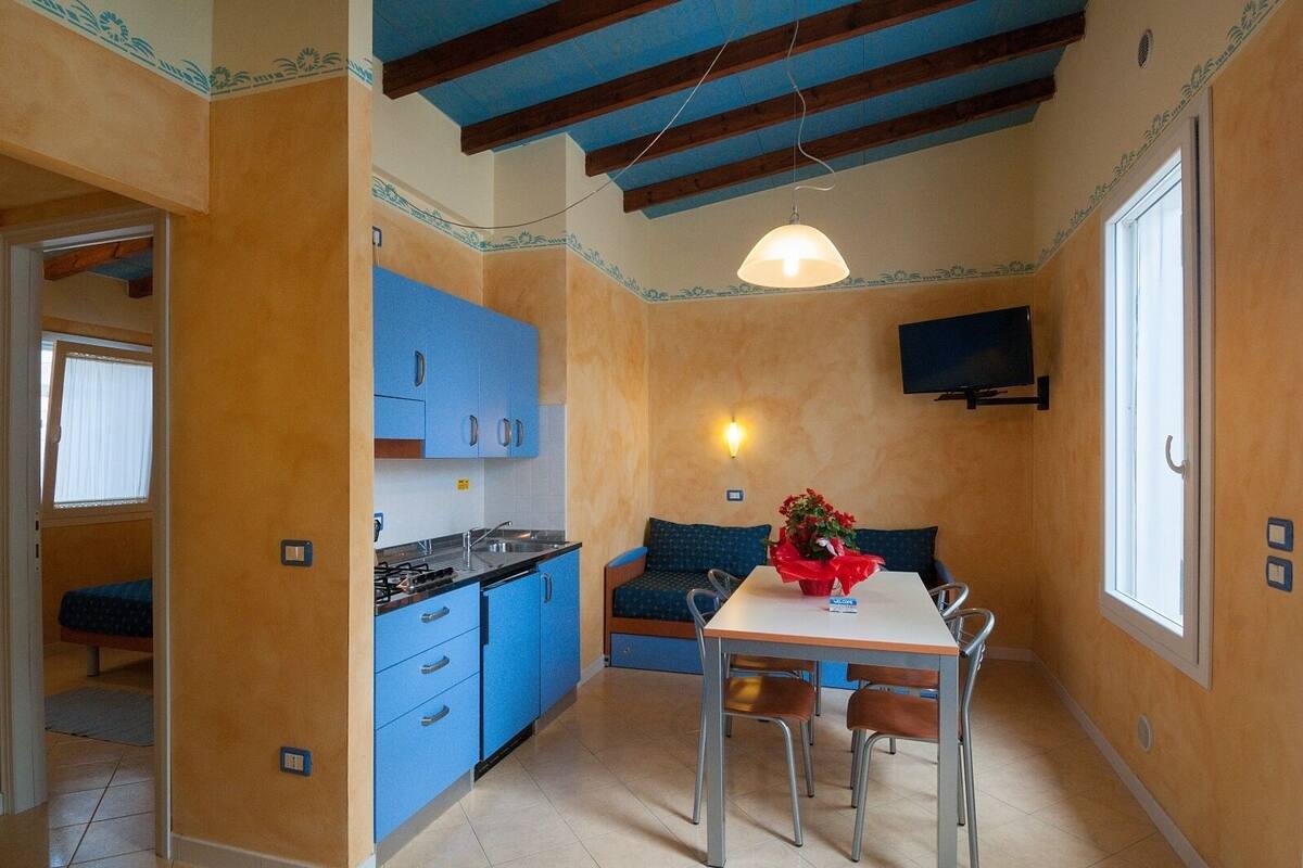 A compact kitchen with blue cabinets, a dining table with chairs, a sofa, and a TV mounted on the wall.