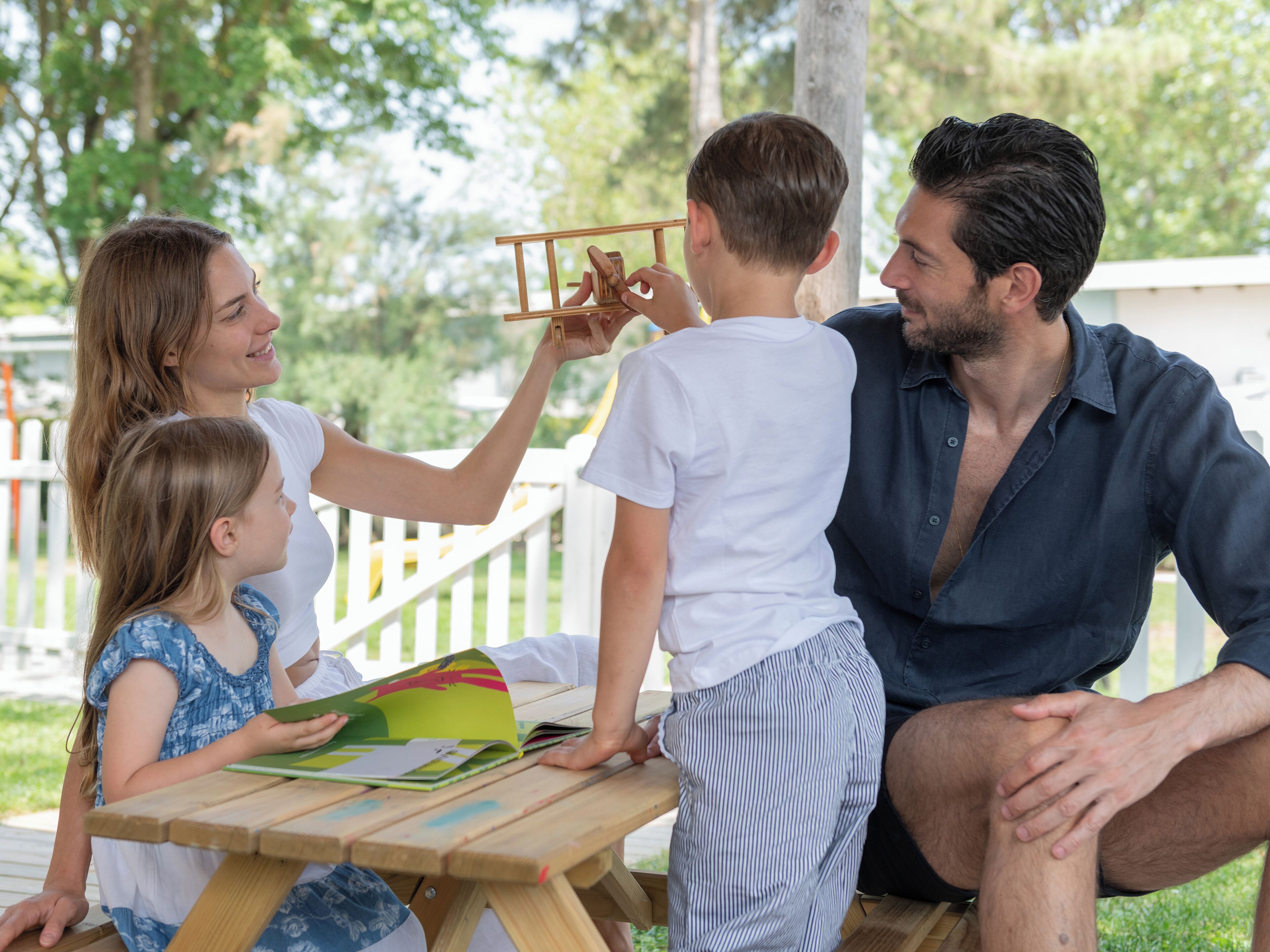 Una familia de cuatro personas en una mesa de picnic de madera al aire libre, con una cerca blanca y follaje de fondo.