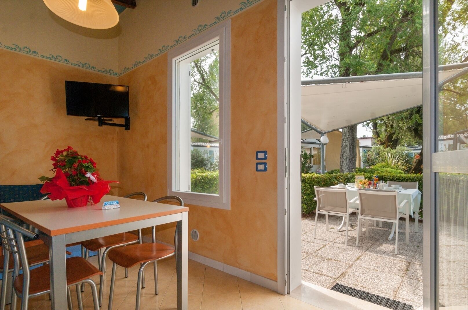 Ein Zimmer mit Tisch und Stühlen, einem an der Wand montierten Fernseher und einer Tür zu einem Außenbereich mit Esstisch.