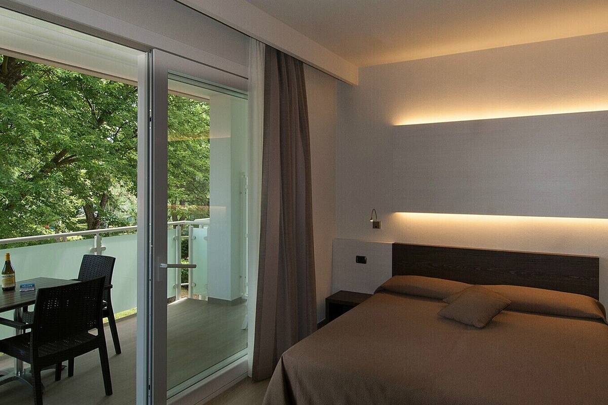 A modern hotel room with a bed, bedside tables, and a balcony with a table and chairs.