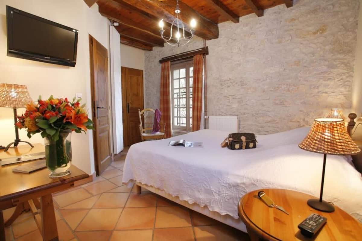 A bedroom with a bed, a TV, a vase of flowers, a lamp, and a suitcase on the nightstand.