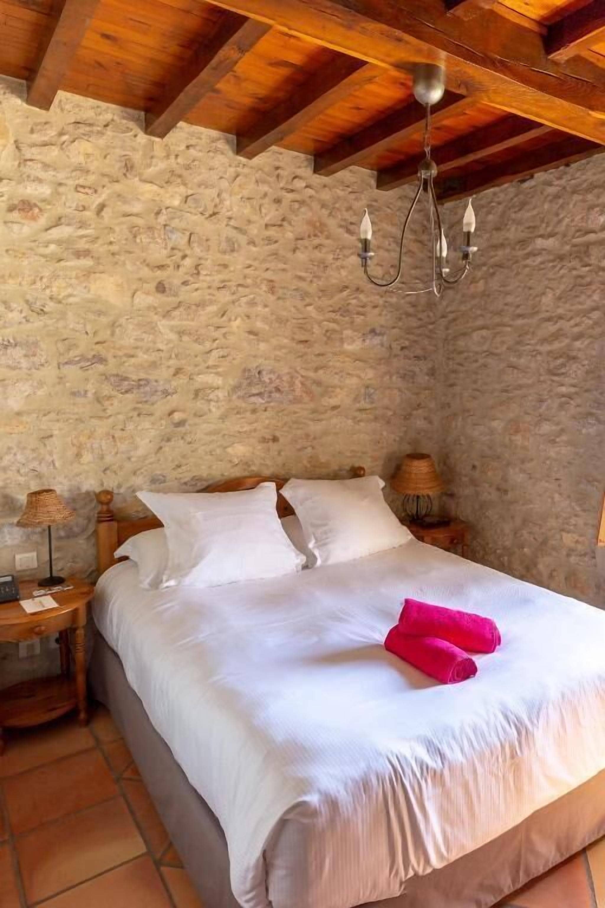 Une chambre avec un mur en pierre, des poutres en bois au plafond, un lit avec du linge de lit blanc, deux serviettes roses et une table de chevet avec une lampe.