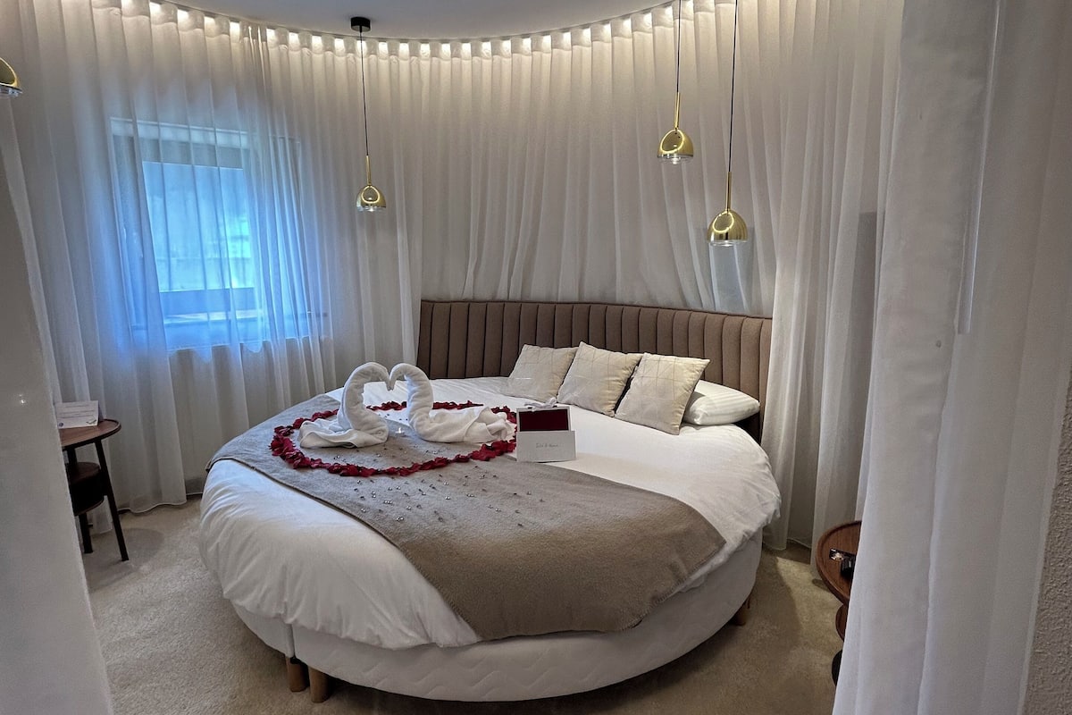 A round bed with a brown headboard, white bedding, and a bouquet of white flowers.