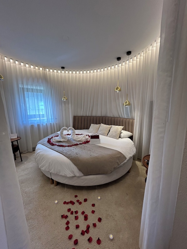 Una cama redonda con cabecera marrón, ropa de cama blanca y un ramo de flores blancas.