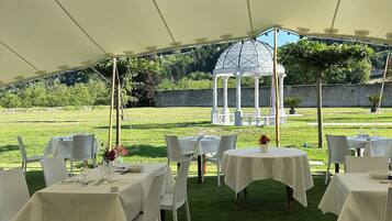 A tented outdoor dining area with tables set for a meal, a gazebo in the background, and a grassy field.
