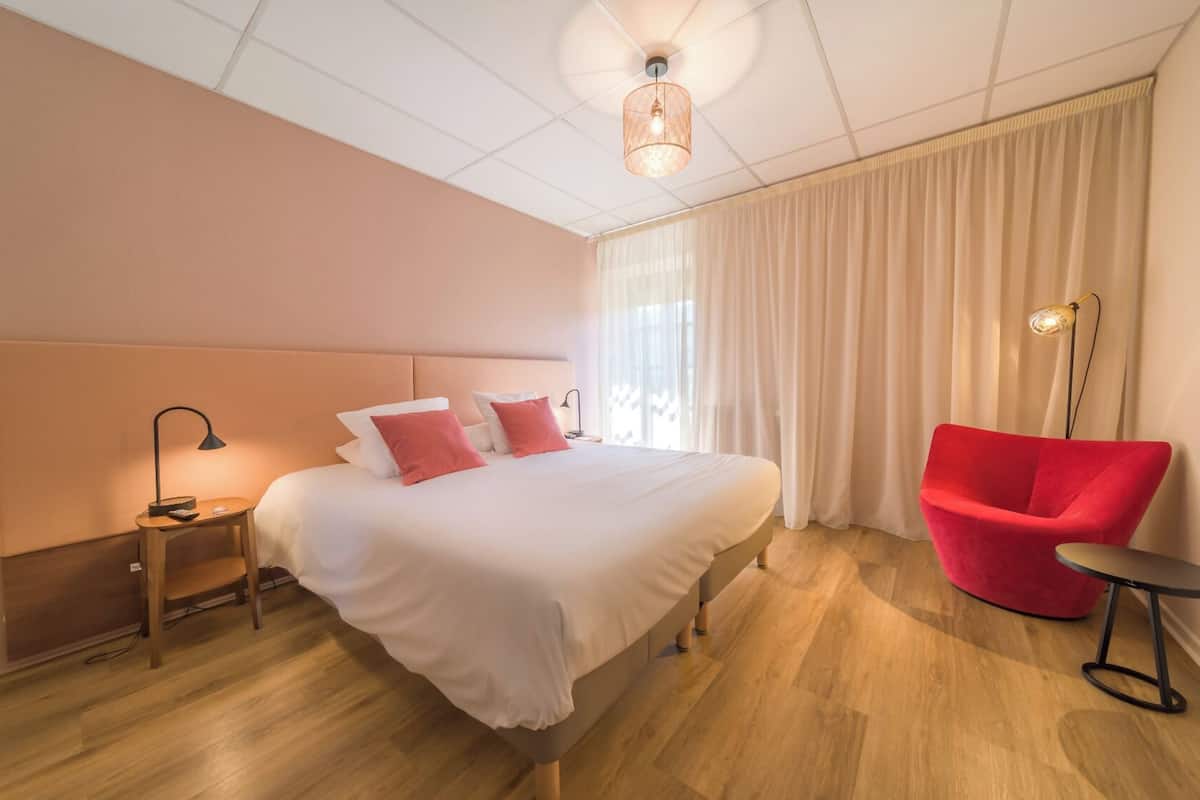 A hotel room with a large bed, a red chair, a bedside table with a lamp, and a ceiling light.
