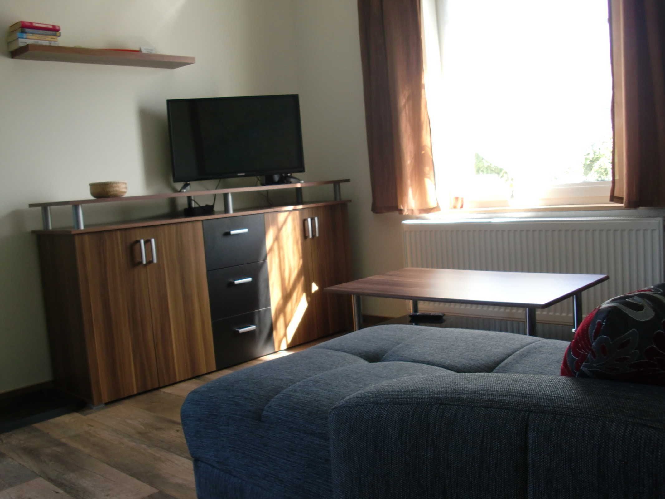 Ein Zimmer mit einer Couch, einem Fernseher auf einer Holzkommode, einem Tisch und einem Fenster mit Vorhängen.