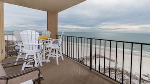 A balcony with white Adirondack chairs and a table, overlooking a beach with a wooden walkway.