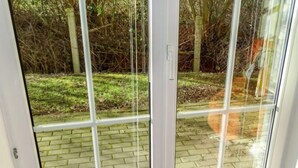 Uitzicht door een glazen deur op een terras van bakstenen en een bebost gebied op de achtergrond.