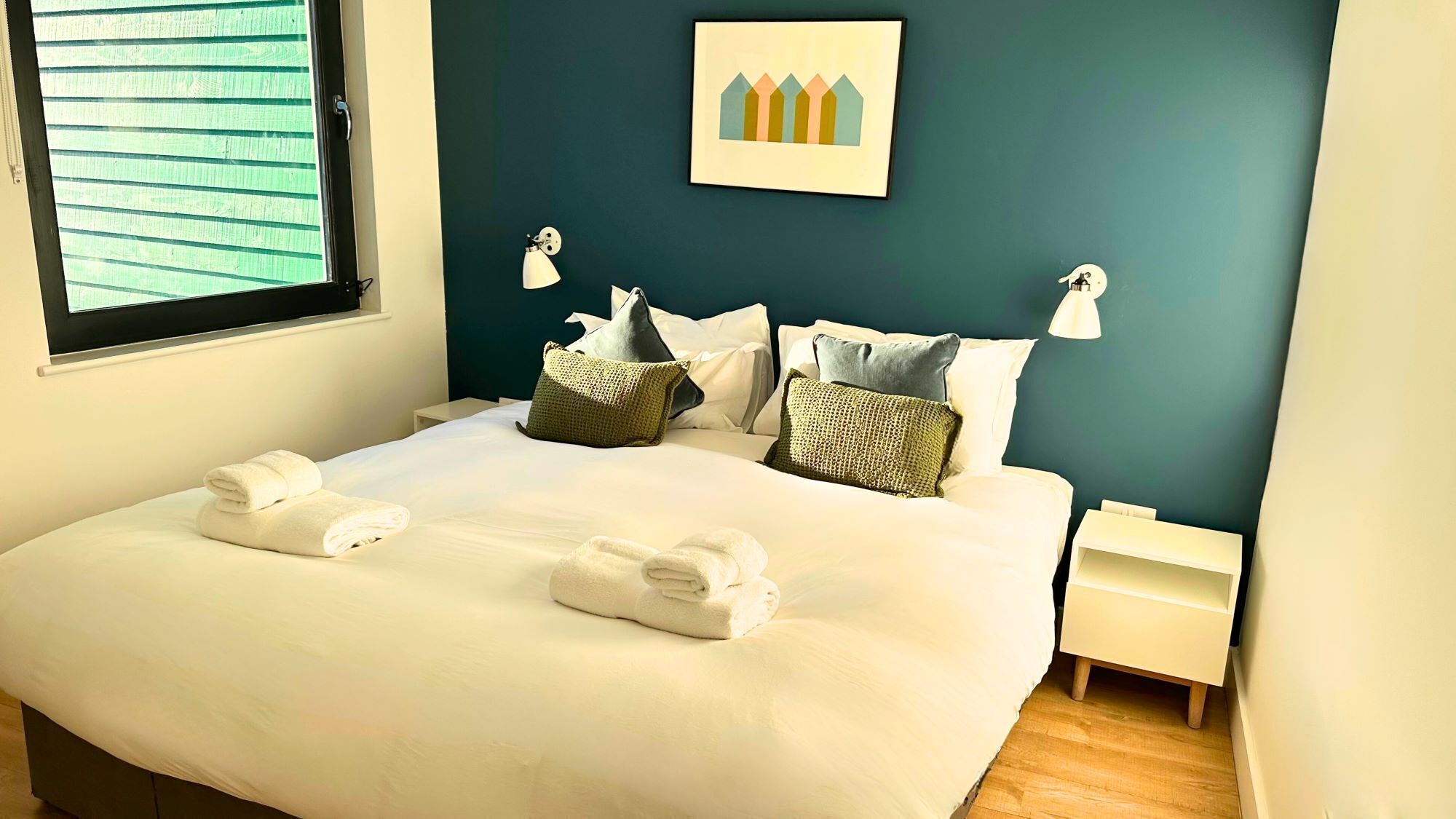 Una cama bien hecha con toallas, una mesita de noche, una lámpara de pared y un cuadro enmarcado sobre la cama.