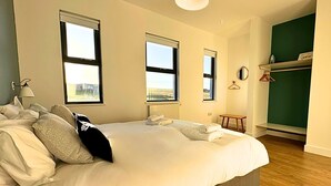 A bedroom with a large bed, two windows, a bedside lamp, a small table, and a green wall. - Kitty Lass - 2 Bed Luxury Apartment (Wick)