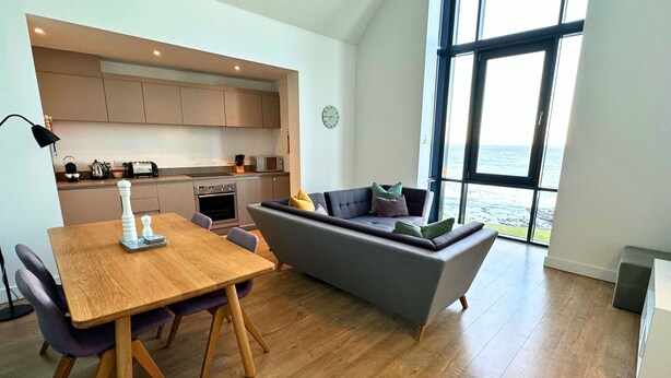 A modern kitchen with built-in appliances, a dining area with a wooden table and chairs, and a grey sofa with cushions. - Kitty Lass - 2 Bed Luxury Apartment (Wick)