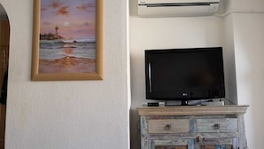 A room with a painting of a lighthouse, an air conditioner, and a television on a wooden cabinet.