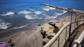 A coastal view with a rocky shoreline, a pier extending into the sea, and a fenced area with beach chairs and umbrellas.
