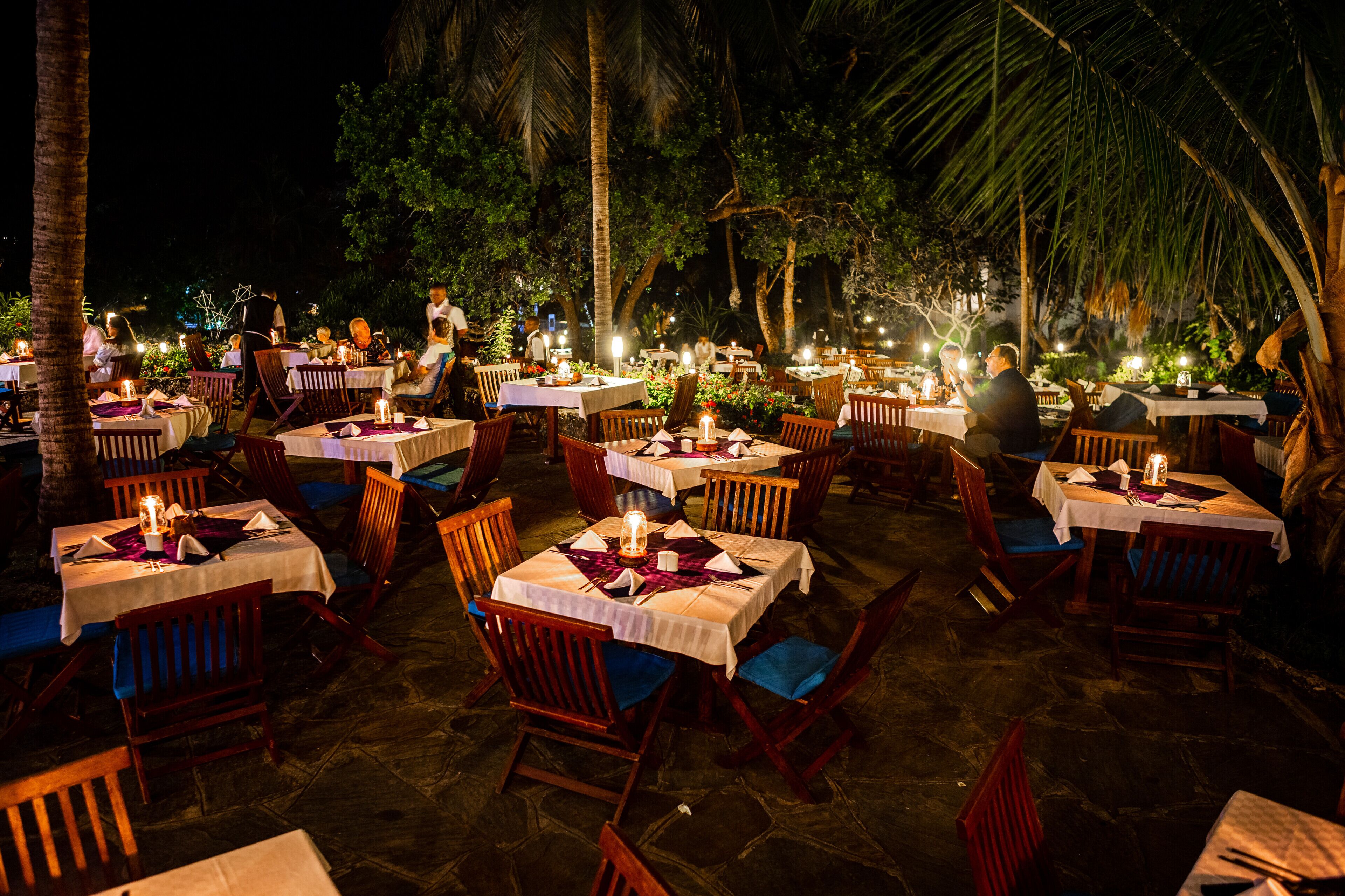 An outdoor dining area with tables set for guests, surrounded by palm trees and under a covered roof.