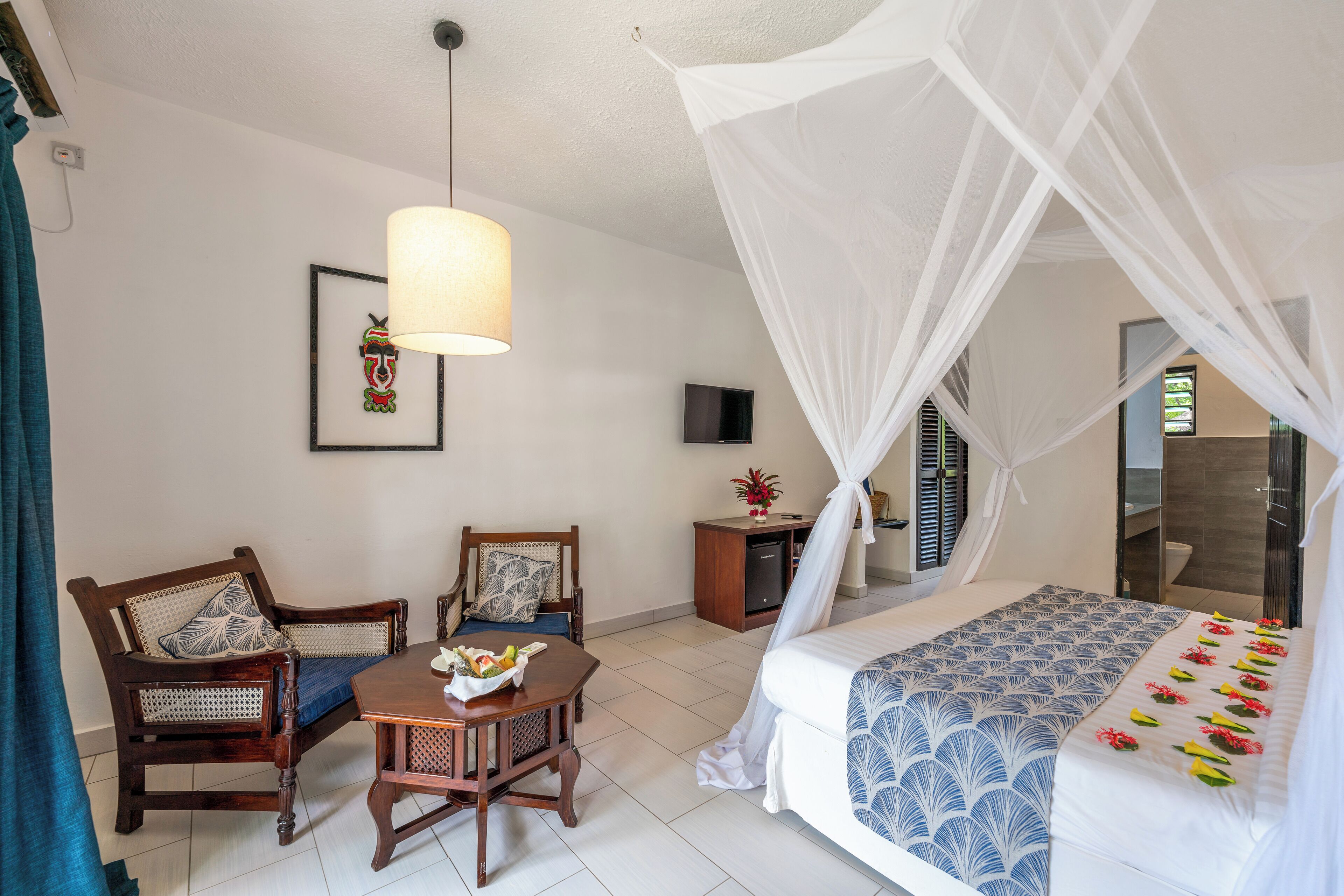 A hotel room with a bed, two chairs, a table with fruit, a TV, and a bedside table with a flower.