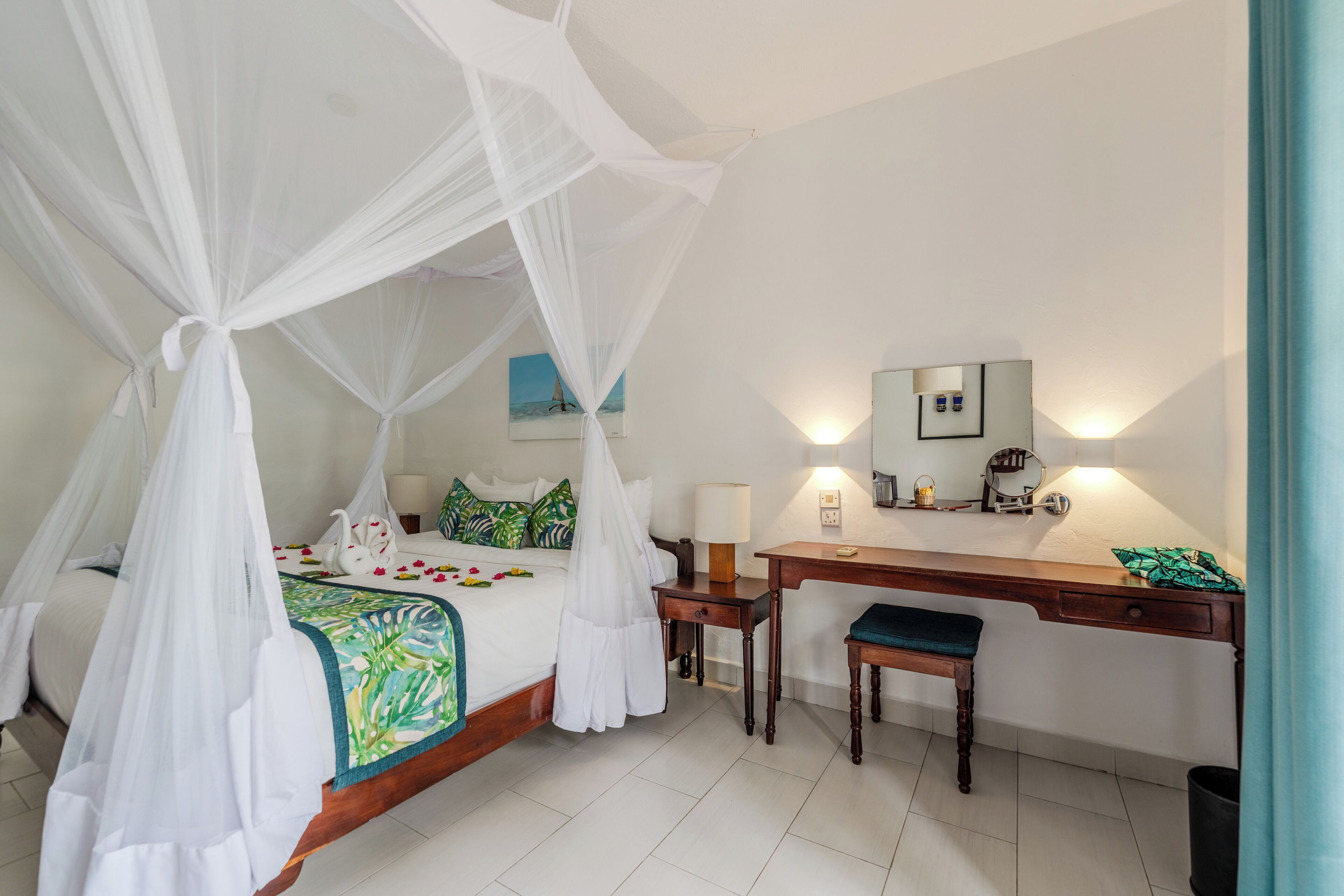 A bedroom with a four-poster bed, a desk with a chair, a bedside table, and a wall-mounted mirror.