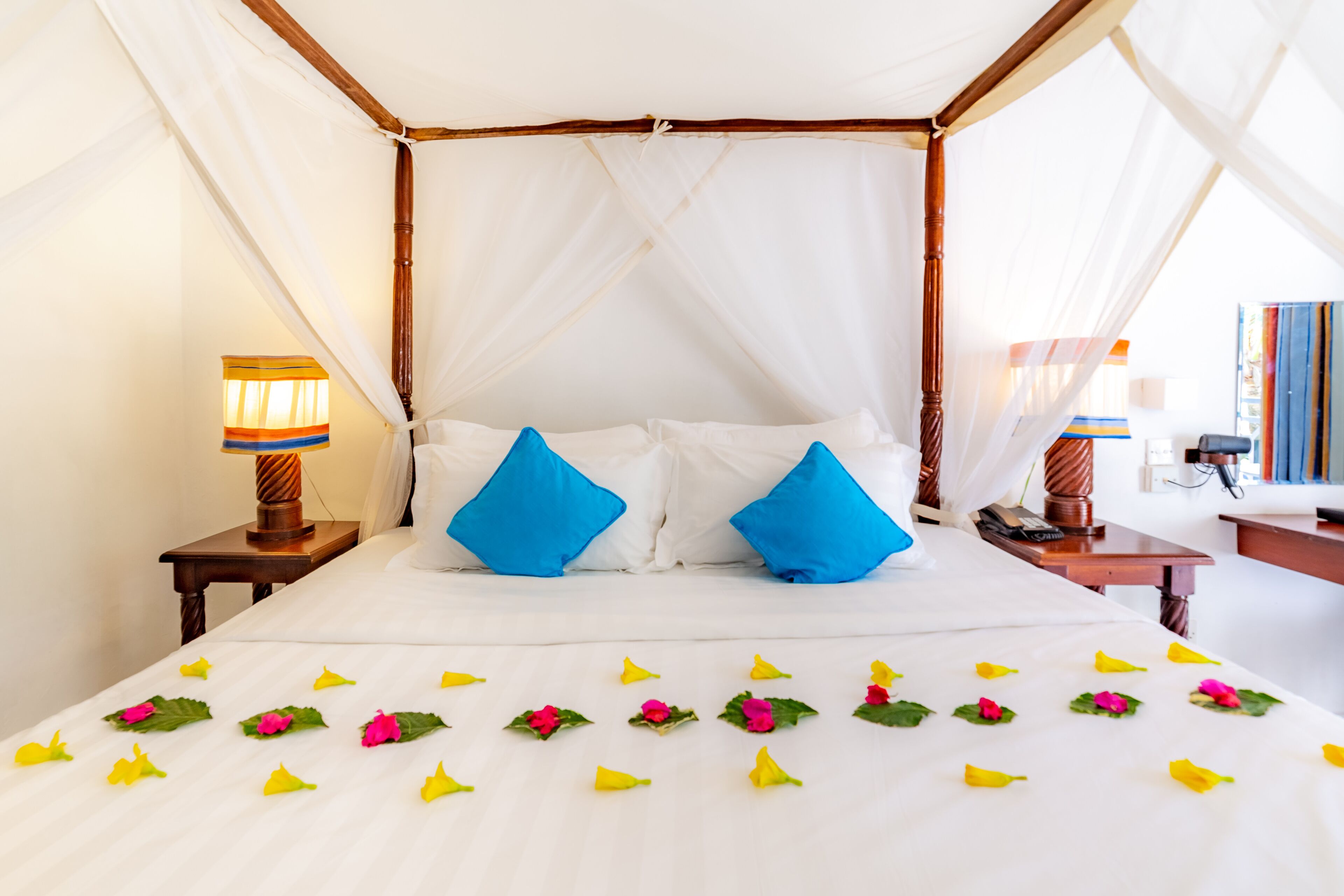 A four-poster bed with white linens and blue pillows, a bedside table with a lamp, and a room with a white canopy.