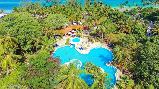 Vista aerea di un resort con una grande piscina circondata da vegetazione lussureggiante e palme, con una spiaggia e l'oceano sullo sfondo.
