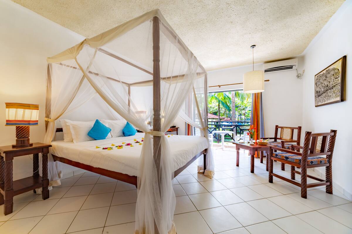 A room with a four-poster bed, a wooden side table with a lamp, a dining table with chairs, and a view of greenery outside.