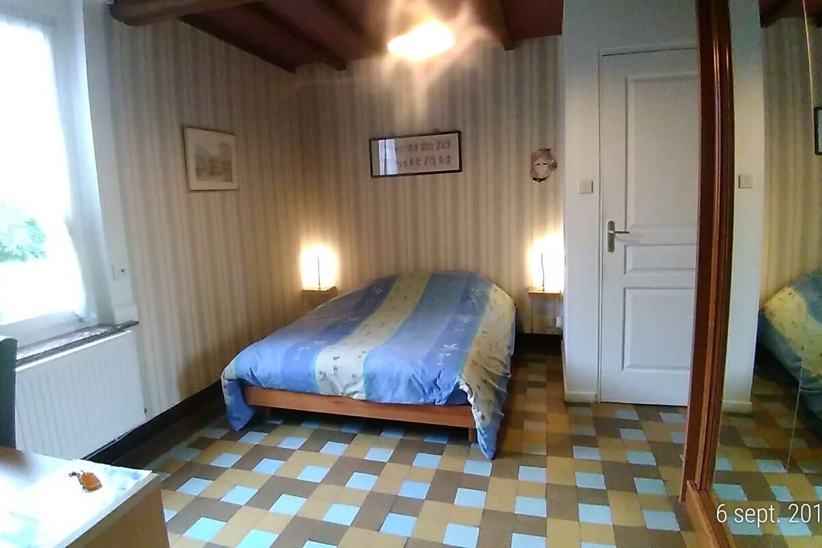 Un dormitorio con suelo a cuadros, cama con colcha a rayas, dos lámparas de noche, un cuadro enmarcado en la pared y una ventana con cortinas.