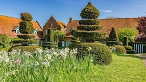 A traditional European-style building with a tiled roof, surrounded by well-maintained hedges and a grassy foreground with flowers.