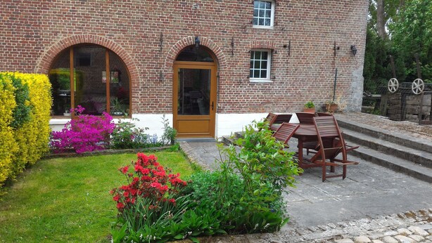 A brick building with a wooden door, arched windows, and a small garden with flowers and a table with chairs.