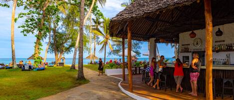 A beachside bar with a thatched roof, palm trees, and a view of the ocean.