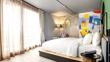 Quarto de hotel moderno com uma cama grande, uma mesa lateral, um abajur e uma pintura abstrata colorida na parede.