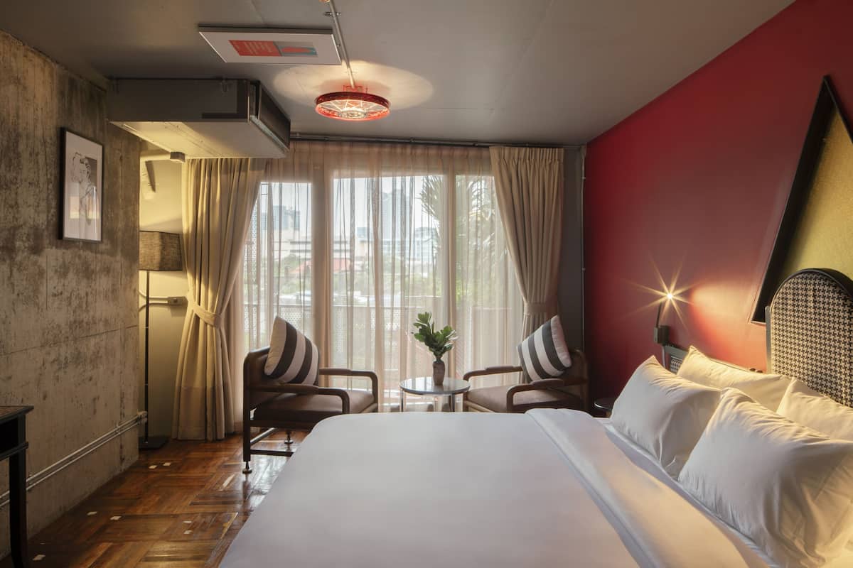 A hotel room with a large bed, two armchairs, a small table with a plant, and a view of the outdoors through large windows.