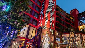 Et moderne hotel med en markant facade i rødt og sort, store vinduer og et muralmaleri af en kvinde på siden.