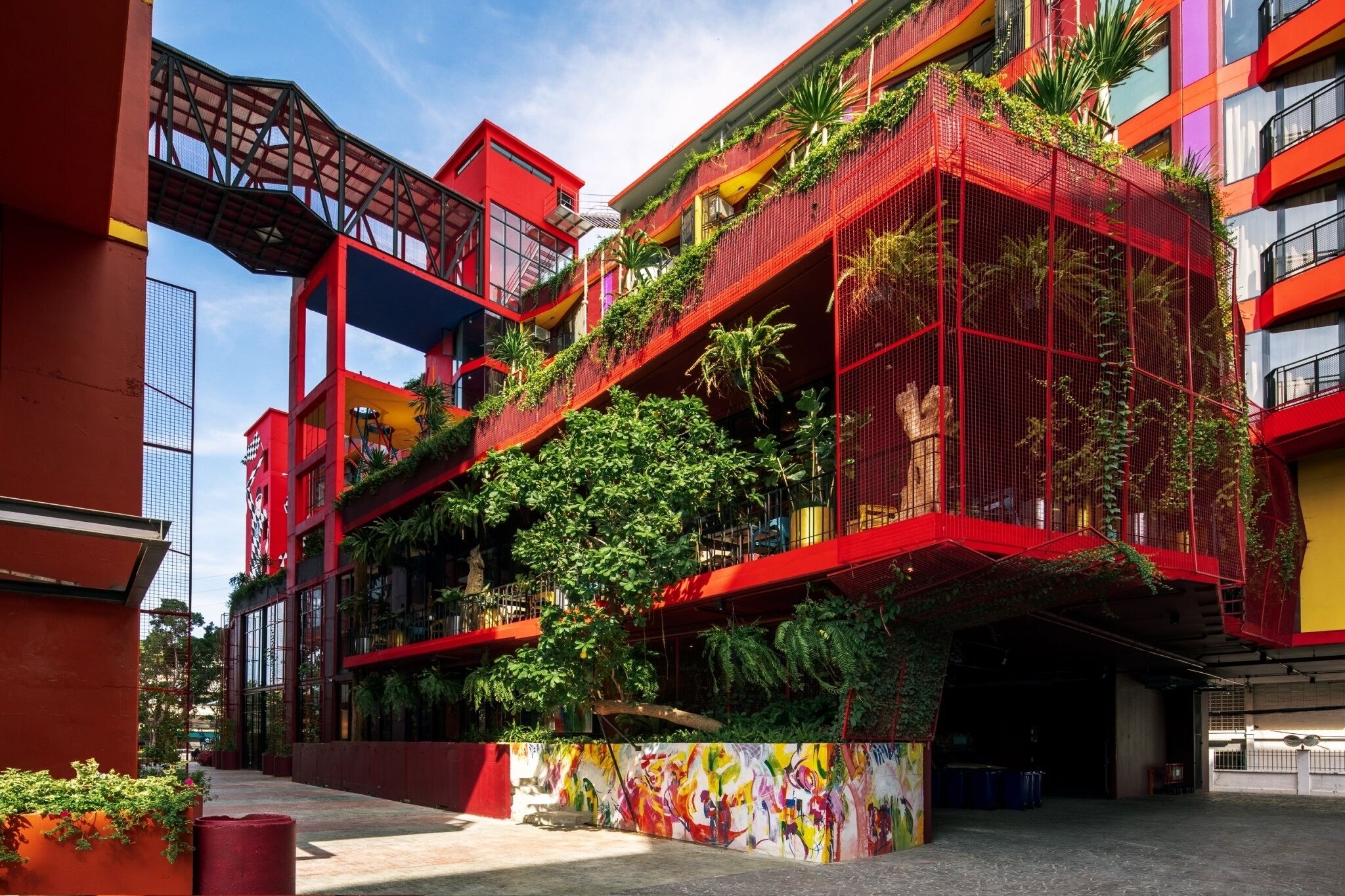 A modern building with red and yellow facades, balconies with greenery, and a colorful mural.