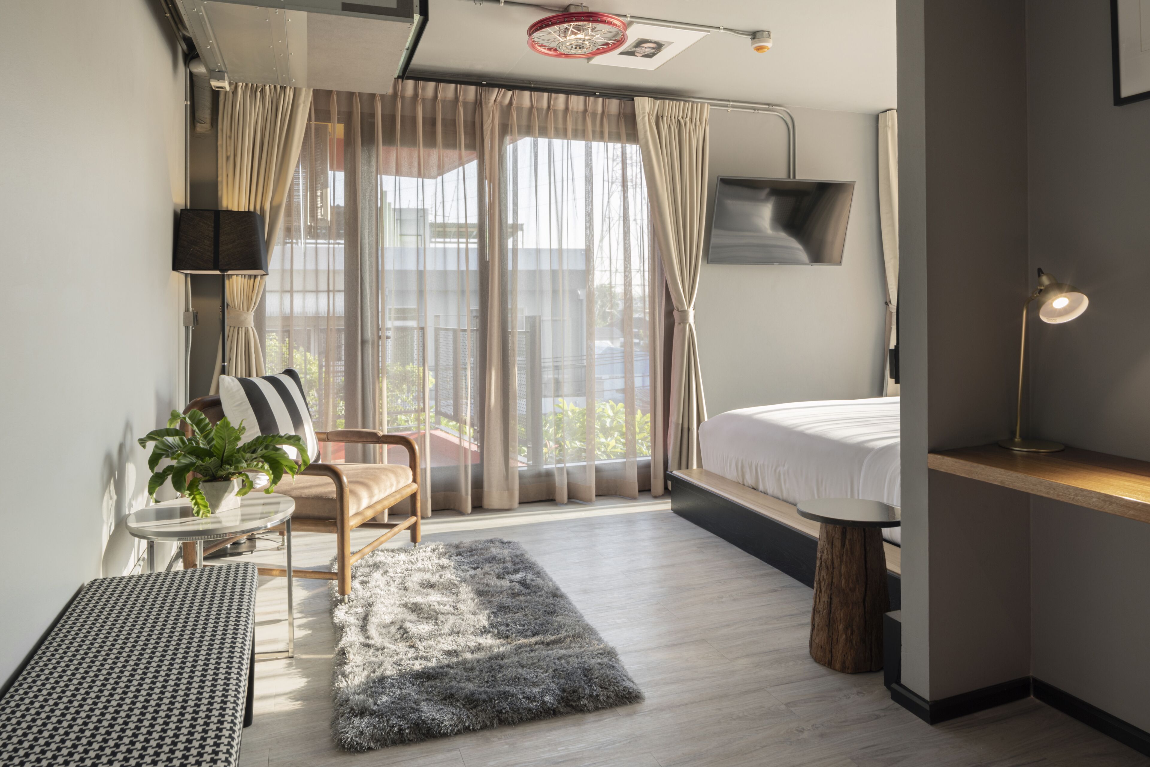 A modern hotel room with a bed, a desk, a chair, a small table, a plant, and a view of the outdoors.