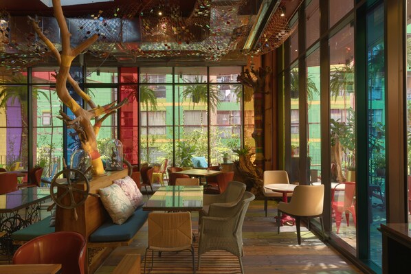 A spacious indoor area with multiple seating arrangements, large windows offering views of greenery, and a decorative tree structure. - MeStyle Museum Hotel (Bangkok)