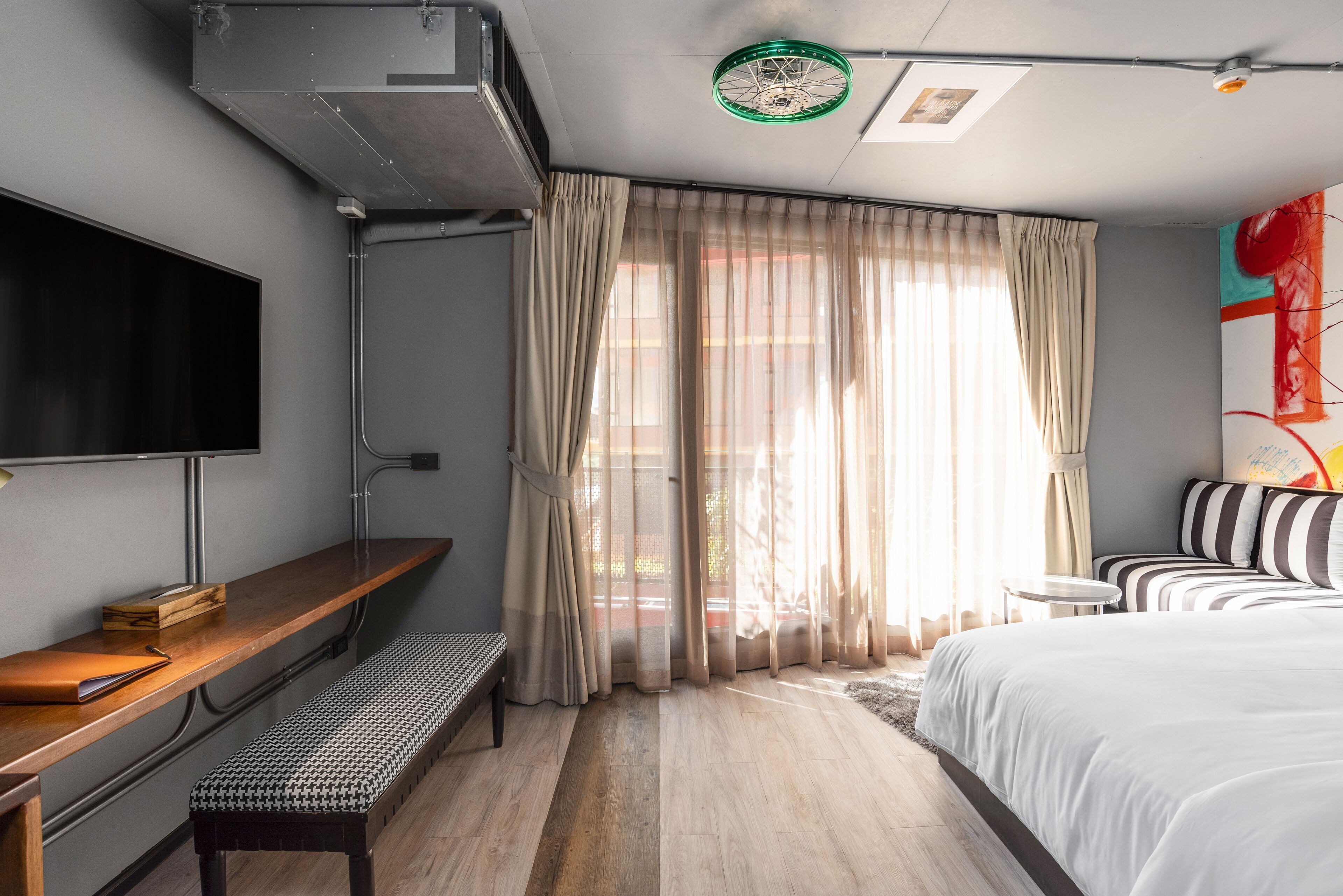 A modern hotel room with a large bed, a bench, a TV, and a window with sheer curtains.