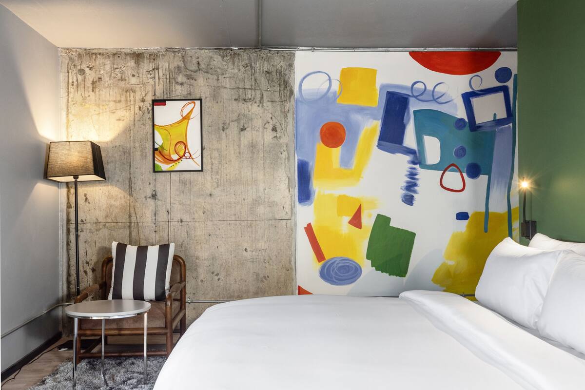 A bedroom with a bed, a chair, a lamp, and a colorful abstract painting on the wall.