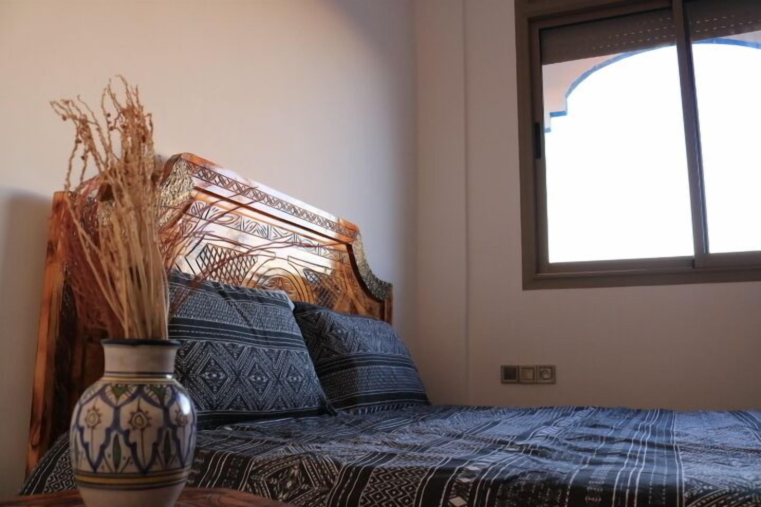 A bed with patterned bedding, a decorative vase with dried plants, and a window with a view.