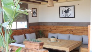 A wooden dining area with a bench and a table, surrounded by potted plants and a wooden wall with a logo. - Azrac Surf Morocco (Aourir)