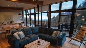 Ein großzügiger Wohnbereich mit einer geräumigen, dunkelblauen Eckcouch, einem Holztisch und Blick ins Freie durch große Fenster.