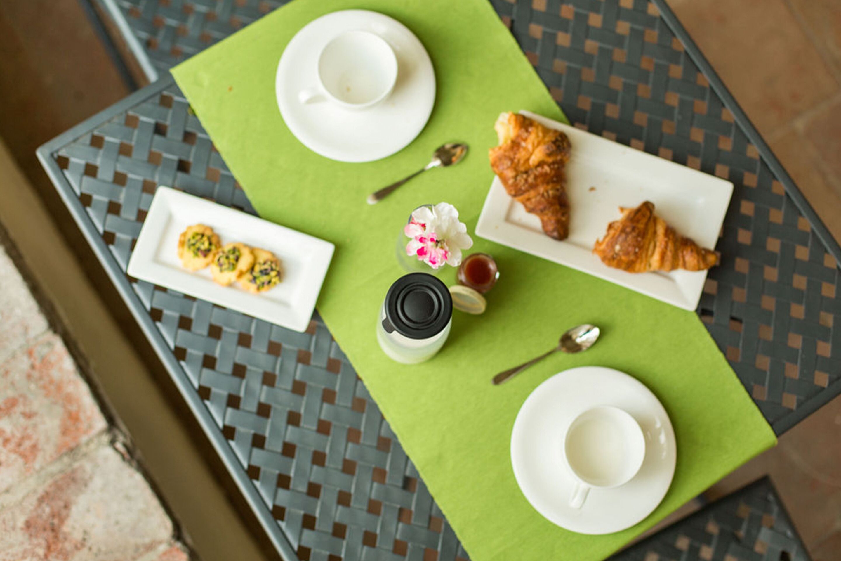 Una mesa con un mantel individual verde, dos platos blancos, un tarro de mermelada, una flor y un croissant.